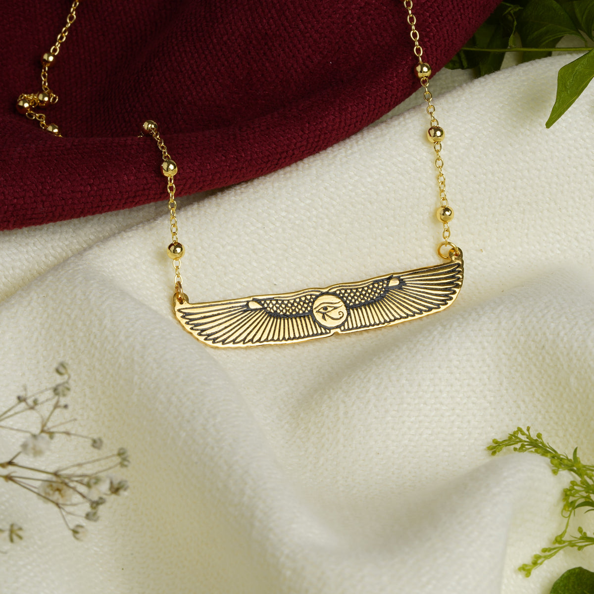 A gold wing pendant necklace with a central circular emblem with eye of Horus in the middle, displayed on draped white fabric with a dark red cloth and green leaves as styling props.